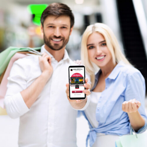 Couple Showing Cellphone Screen Standing In Shopping Center, Mockup Shopping App. Smiling Couple Showing Blank Cellphone Screen Standing In Mall Center. Mockup, Shallow Depth